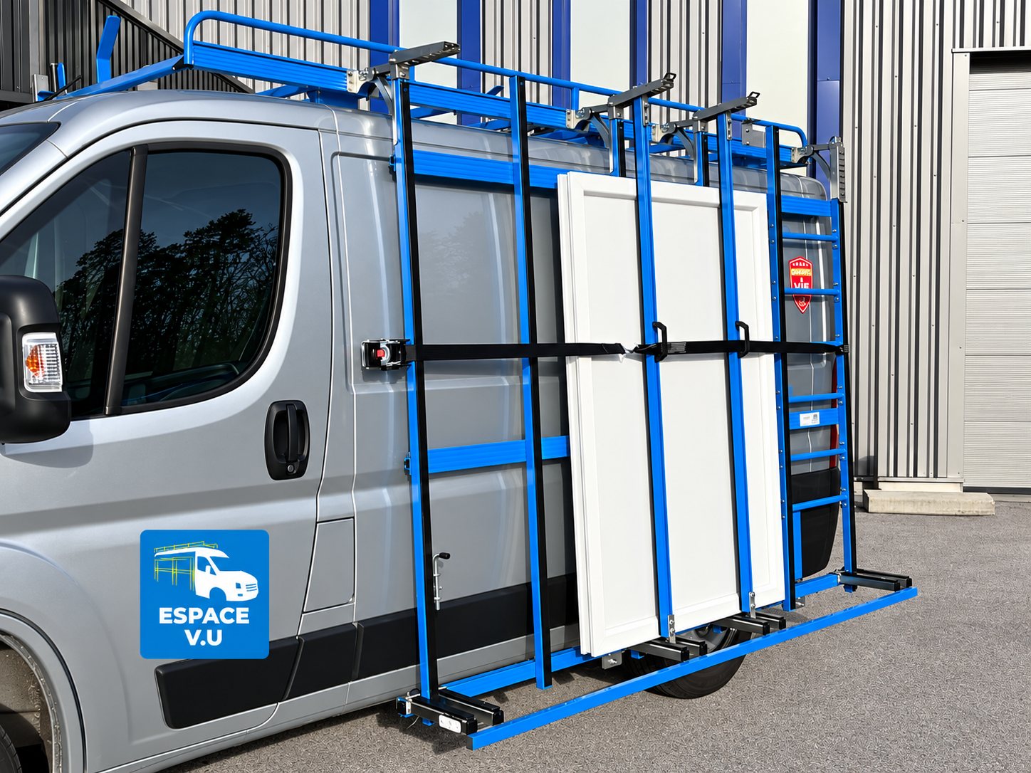 Un fourgon utilitaire gris est équipé sur le côté d'un porte-vitrage bleu avec sangles, qui maintient un panneau de porte blanc devant un bâtiment à bardage métallique.